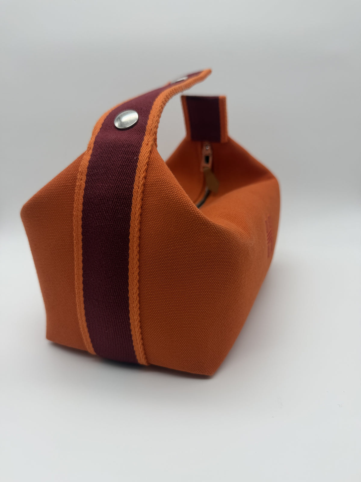 Three-quarter view showing the bordeaux webbing strap edged in orange and a palladium snap stud